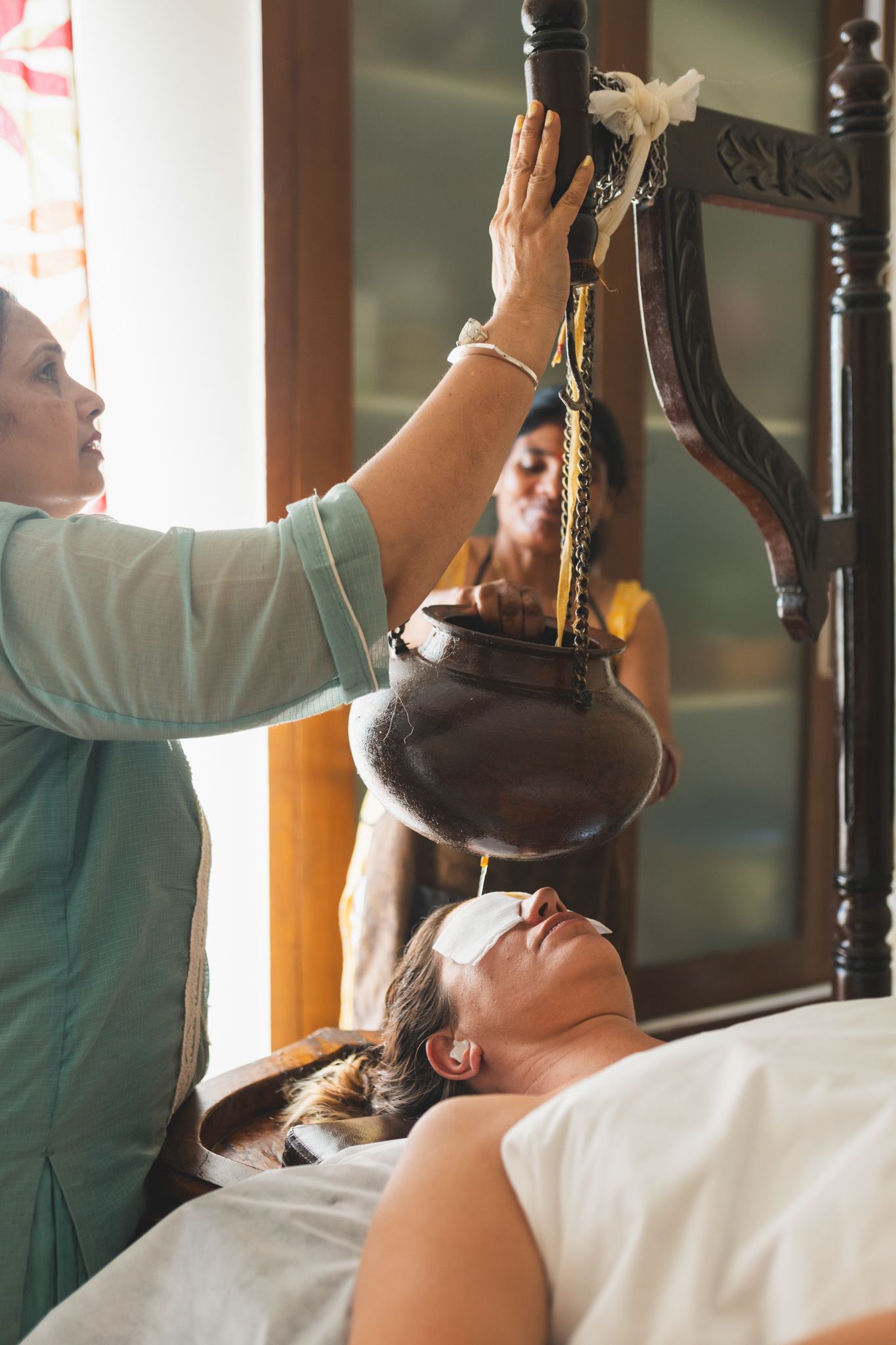 A therapist in a pale green kurta adjusts a Shirodhara pot hanging from a carved wooden frame above a woman lying on a treatment table with pads over her eyes